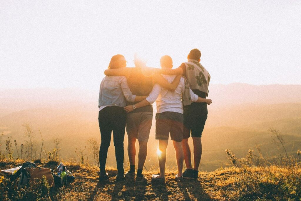 Four friends stand with their backs to the camera, their arms around each other's shoulders and waists, staring at a beautiful sunset that is cresting over a hilly background, representing cigna mental health rehab coverage accepted at Transformations Mending Fences making treatment accessible